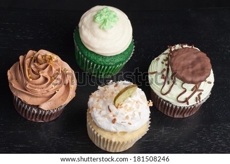 Coconut Key Lime Cupcake, Pot of Gold Chocolate Caramel Cupcake, Irish Mint Chocolate Cupcake, and Green Velvet Cupcake topped with cream cheese frosting on a black background