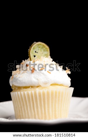 Key Lime Coconut Cupcake - coconut cake with a key lime filing topped with a 7 minute frosting, toasted coconut, and a slice of candied key lime isolated on black on a vintage antique plate