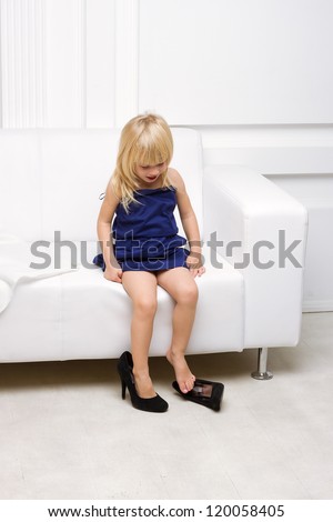 Little girl 3 years old sitting on a white sofa in her mother\'s dress and shoes high heels