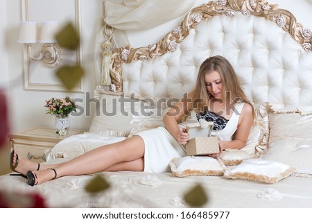 Stylish blond woman in black dress  in hotel apartment. Sexy girl gifts relaxing on bed at home luxury interior