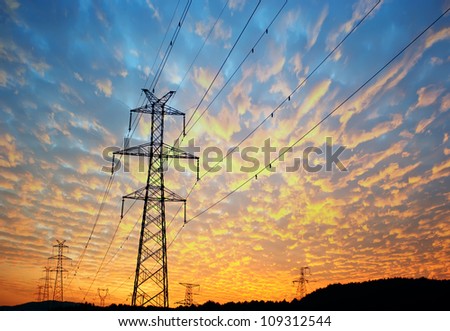 Electricity pylons, power lines and trees silhouetted against a cloudy sky at sunset.