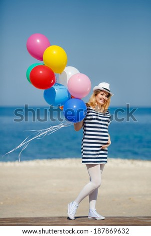 Portrait of beautiful pregnant woman in sunglasses and white hat with bright colorful balloons on sea shore background