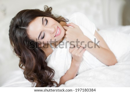 Beautiful young brunette woman with attractive smile laying on white cozy bed embracing pillow - indoors. Portrait of woman in modern bedroom.