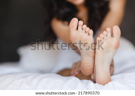 Beautiful feet of a young woman lying in bed close up. Woman Holding her Tired Feet in Hand Sitting on Bed with White Sheets. eet of a woman sleeping on the white linen at home