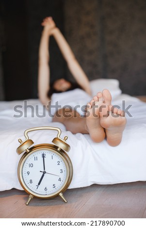 A clock and the feet of a sleeping woman. Early morning.sleeping young female disturbed by alarm clock early morning on bed