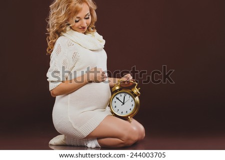 portrait of pregnant woman with clock in her hands on brown background. pregnant woman holding alarm clock. blonde young woman