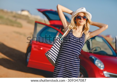 Summer vacation. Car trip. Travelling. Car travel. By the sea. Beautiful blonde woman standing with red small car on the background. Sea style. Summer vacation car road trip freedom concept.
