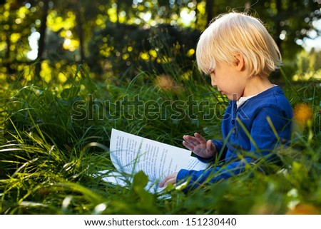 little cute blond boy reading book on the grass outdoor