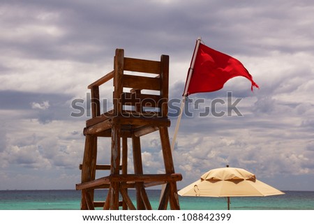 Abandoned lifeguard chair at a Caribbean beach, with the horizon over the ocean in the background and a red flag showing unsafe conditions, under a cloudy sky.
