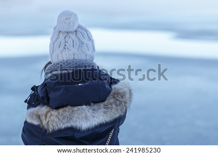 Back of a young woman in think winter clothes as she looks towards a frozen lake.
