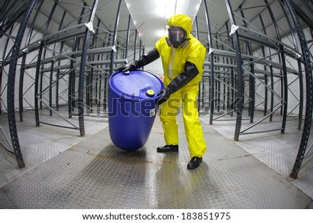 Worker in protective uniform,mask,gloves and boots rolling barrel of chemicals in empty storehouse - fish eye lens
