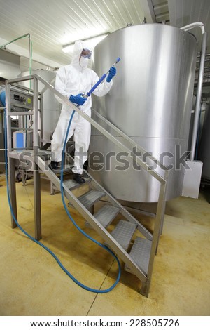 worker in white protective uniform,mask,gloves with high pressure washer on stairs at large industrial process tank preparing to cleaning