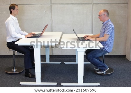 two men  coworking in correct sitting posture on pneumatic leaning seats  with laptops  at electric height adjustable desks in office