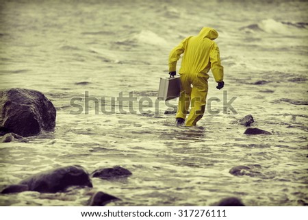 technician  in protective suit with silver case  walking in water at rocky beach - back view