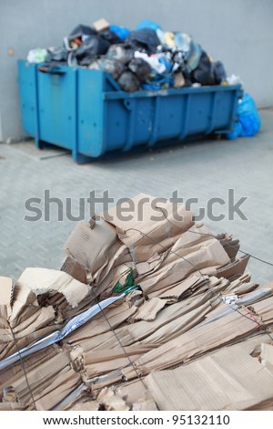 tied,segregated cardboard at the front and waste containers with nonsorted wasted in bags at the back - out of focus