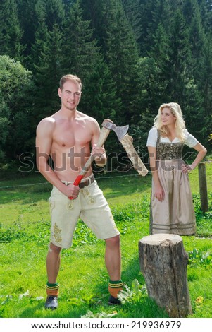 Bavarian couple in love in fashionable dress clothing for firewood work on a summer pasture in the mountains / Bavarian couple in love chopping wood in fashionable dress clothing