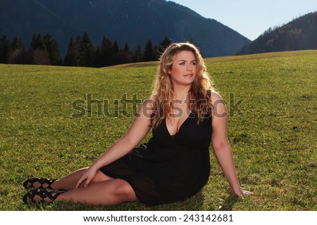 Blonde elegant woman with oversize half lying in black dress and wide neckline on a meadow in the Alps / Elegant woman with oversize half lying on a meadow in the Alps