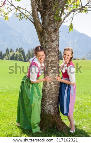 Two smiling women in dirndl hold on to a tree in a meadow in the Bavarian Alps.