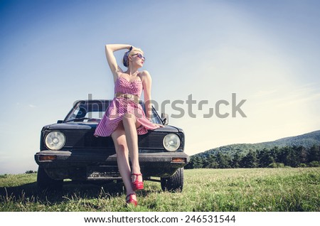Sexy dressed vintage female model posing next to retro car on a field