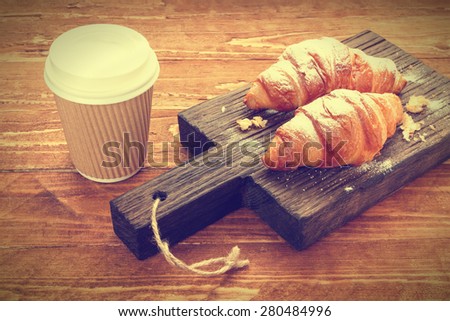 Two croissants and coffee-to-go cup on rustic cutting board. Vintage effect