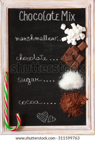 Hot chocolate mix on old chalk blackboard. Sweet marshmallow, chocolate bars, sugar, cocoa powder and peppermint candy cane for cooking winter holiday drink.