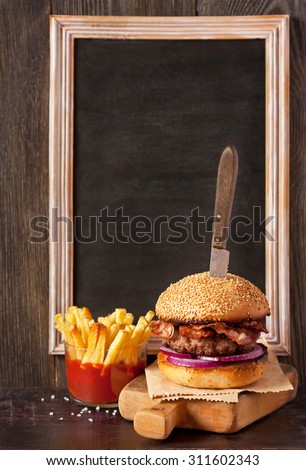 Brutal bacon hamburger served with old knife on wooden cutting board, fried potatoes and chalk blackboard for text.