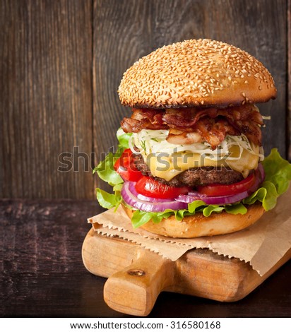 Big delicious cheeseburger stacked high with a bacon, beef patty, coleslaw, cheese, lettuce, red onion and tomato on sesame seed bun served on wooden cutting board.