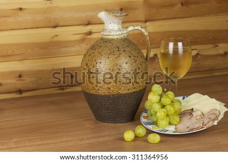 Old clay jug and a glass of wine on a wooden table. White wine and snacks. Ham, cheese and grapes to eat. Relax with wine and good food. Place for your text. Wooden blurred background.