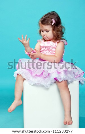 Little, dark-haired baby girl getting ready for the celebration and dressing up in jewelry. Baby dressed in a pink fluffy dress. Isolated on blue background.