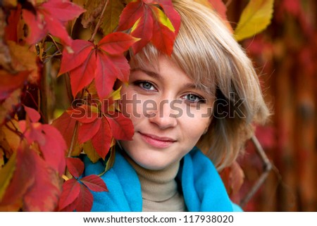 Beautiful face of a young gray-eyed blonde woman near a tree with red autumn leaves.