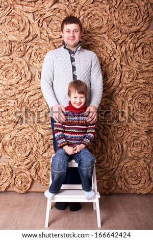 Father and son. Son sitting on a stool, his father standing behind him and hugging him.