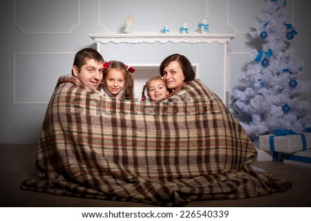 Happy family, mother, father and two kids sitting near the fireplace wrapped in a plaid.