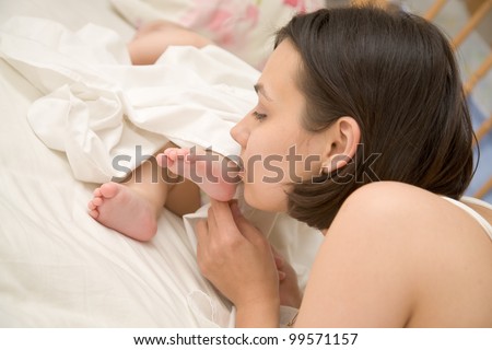 Pretty young mother kissing the tiny feet of her newborn baby who is sleeping in bed