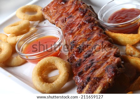 American ribs on white plate with baked potatoes and onion rings - with two red chili sauce on the side