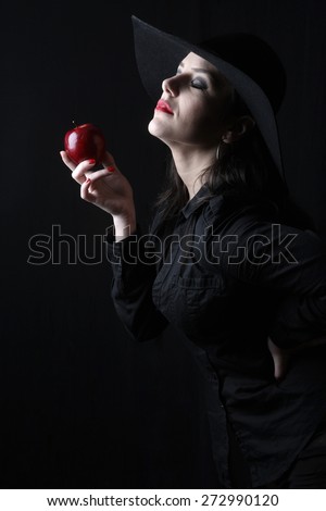 beautiful young brunette woman lady posing in a studio on a black background with old style hat biting a red apple