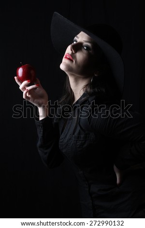 beautiful young brunette woman lady posing in a studio on a black background with old style hat biting a red apple