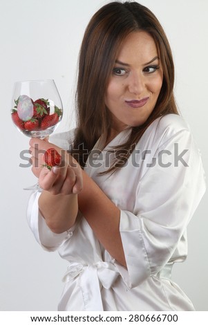 white wine glass of crystal full of fresh organic red nature strawberries - a beautiful young smiling brunette with large breasts in a white sexy silk peignoir