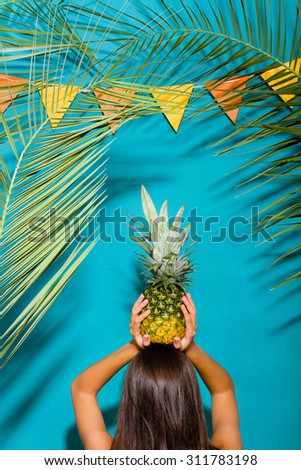 Young woman from the back holding a pineapple above her head. Summer background with palm tree foliage and garland