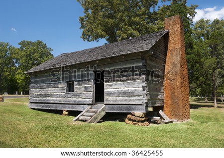 The exterior of a log cabin on a stone foundation and a clay stucco chimney.