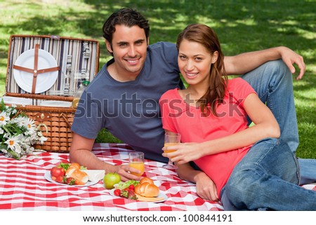 Two friends holding glasses while lying next to each other on a blanket with a picnic basket and food