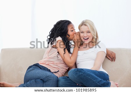 A young woman sitting on a couch is whispering into her friends ear
