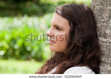 Young relaxed woman looking ahead while leaning against a tree in a public garden