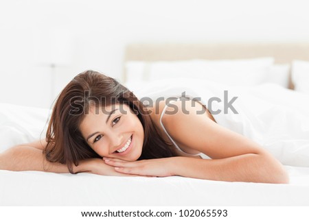 A woman lying at the end of the bed looking forward while smiling and her head resting on both of her hands.