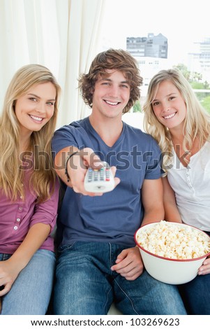 Three smiling friends with a bowl of popcorn and a tv remote looking at the camera