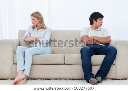 A angry couple sit at the two ends of the couch with their arms folded looking away