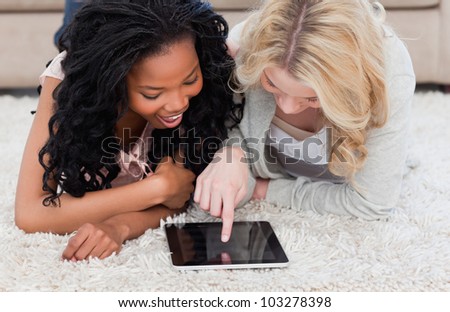 Two women lying on carpet are looking at a tablet in front of them