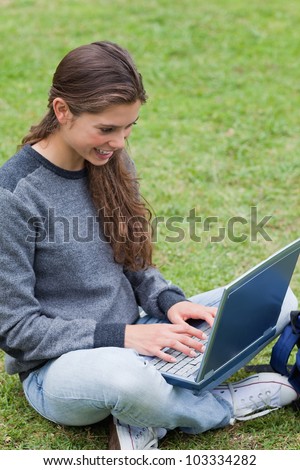 Smiling student typing on her laptop while sitting cross-legged on the grass in a park