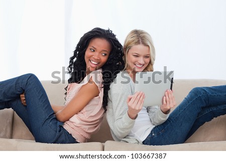 Two women are sitting back to back on a couch and are looking at a tablet
