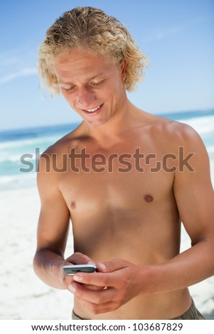 Smiling blonde man standing upright while sending a text with his mobile phone