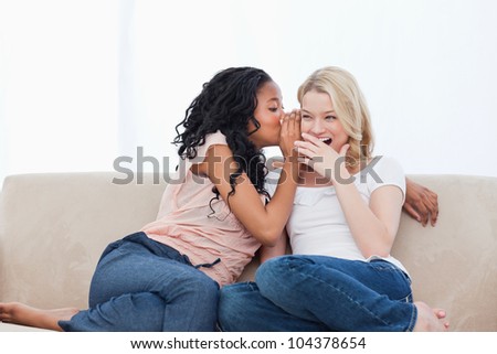 A woman sitting on a couch is whispering into her shocked friends ear